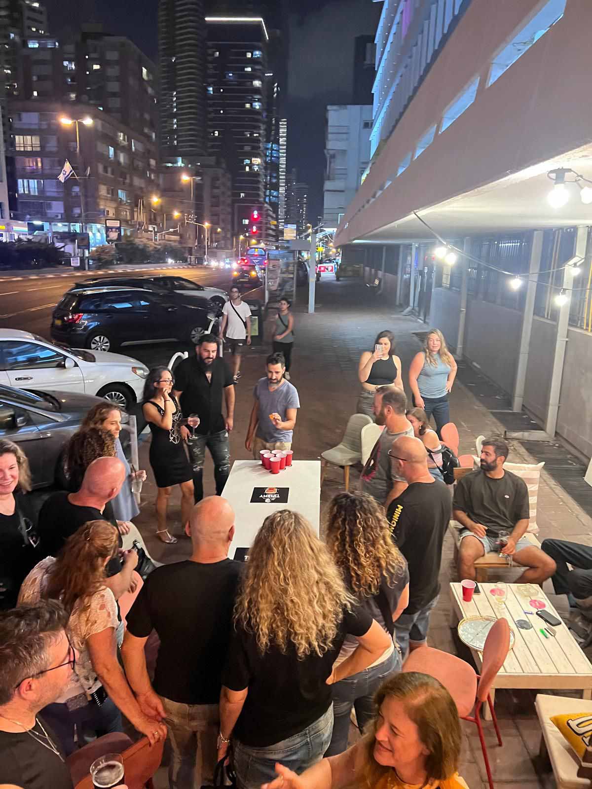 אורחים מסביב לשולחנות הבר בחוץ על המדרכה, רובם עומדים ועל השולחנות כוסות אדומים. בצד שמאל מכוניות חונות בניצב למדרכה, וברקע ביניינים. ברקע מימין חלונות הבר