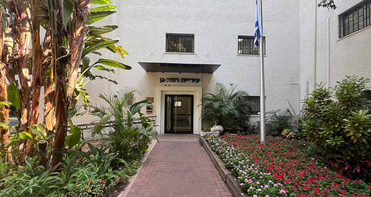 הכניסה לעיריית רמת גן, דלת זכוכית עם מסגרת שחורה, מבט מרחוק. תורן עם דגל המדינה בצד ימין, ועצים בצד שמאל