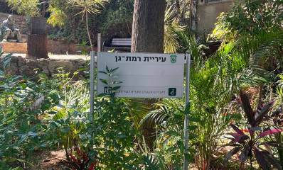 שלט של עיריית רמת גן: מעברים מונגשים ומעלית לשירותכם, וברקע גינה ובה פסל של אדם יושב