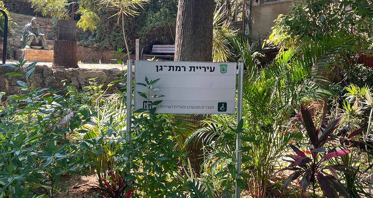 שלט של עיריית רמת גן: מעברים מונגשים ומעלית לשירותכם, וברקע גינה ובה פסל של אדם יושב