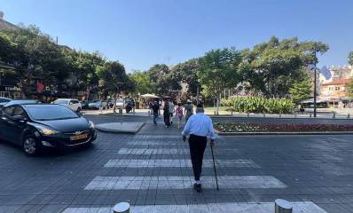מעבר חצייה בשדרה, מכונית שחורה עצרה לקשיש לחצות, האדם נעזר במקל הליכה וחוצה עם גבו אלינו