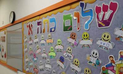 לוח על קיר של כיתה, כותרת: שלום כיתה א', ושמות התלמידים על הלוח על גבי ציור של פרצופים חייכנים ושעון מעל הלוח