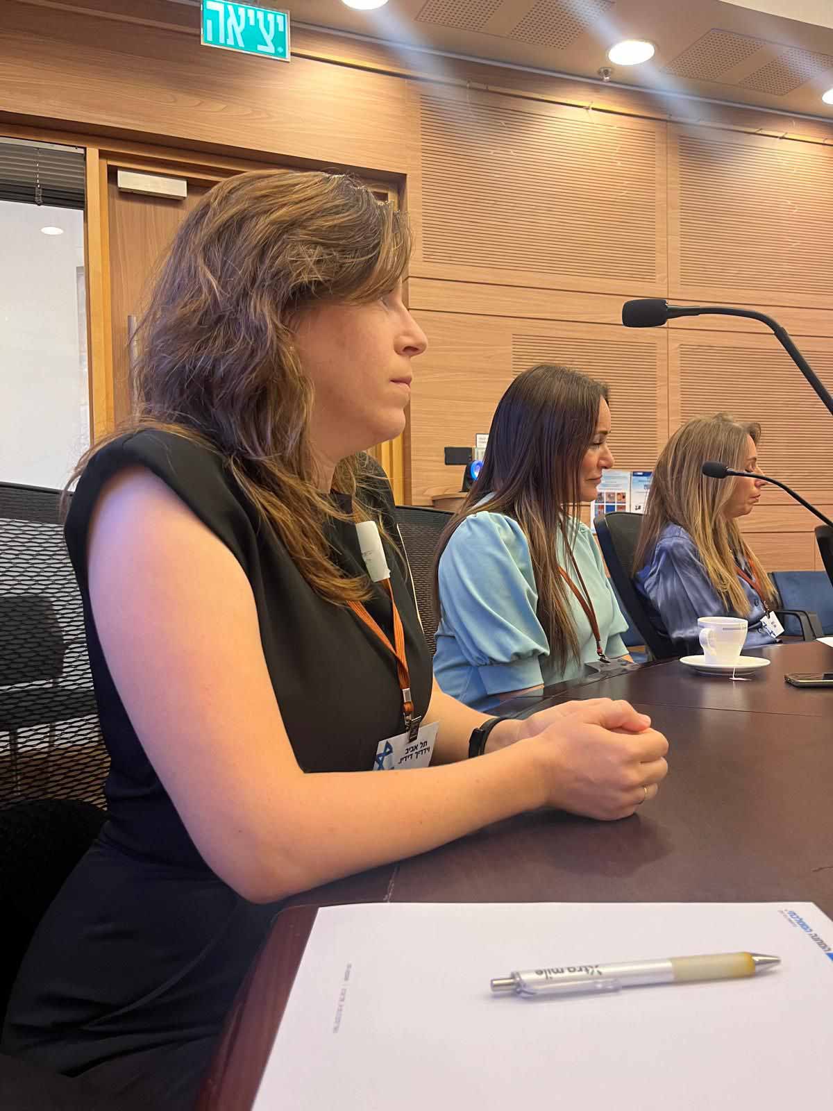 חברת מועצת העיר רמת גן וידריך דידיו, בשמלה שחורה ושיער ארוך, יושבת ליד שולחן הישיבות של העיר. על צוארה בסרט כתום תלוי כרטיס. משמאלה יושבות שתי חברות מועצה נוספות.