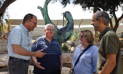 שלושה מתושבי גבעתיים עם ראש העיר רן קוניק עומדים ומשוחחים בגינה ציבורית עם פסל ירוק מאחוריהם, עצים וסלעים בגינה..
