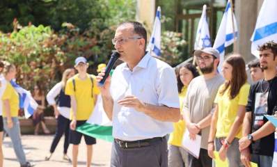 ראש העיר גבעתיים עם מיקרופון ביד עם התלמידים מגבעתיים המפגינים בכיכר החטופים בתל אביב. חלק מהתלמידים בחולצות צהובות. בצד ימין דגלי המדינה וברקע שיחים ועצים.