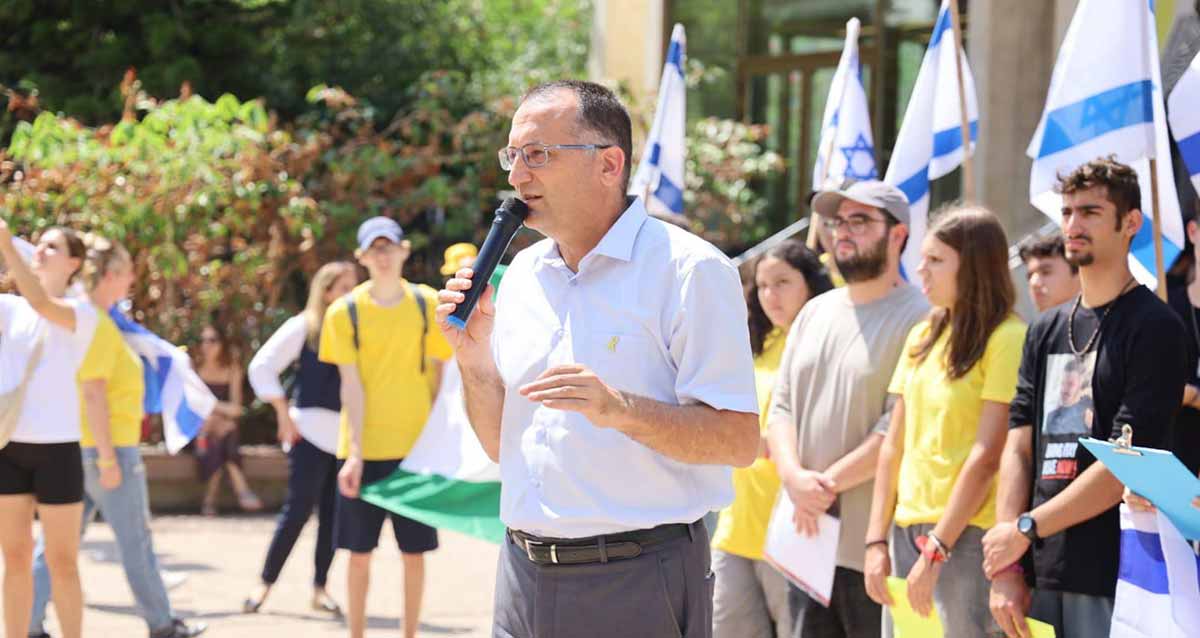 ראש העיר גבעתיים עם מיקרופון ביד עם התלמידים מגבעתיים המפגינים בכיכר החטופים בתל אביב. חלק מהתלמידים בחולצות צהובות. בצד ימין דגלי המדינה וברקע שיחים ועצים.