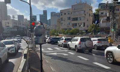 אכיפה בצמתים של שכונת חרוזים. צומת רחובות. במרכז מצלמה על עמוד ולפניה רמזור. מימין ומשמאל מכוניות פרטיות בשני המסלולים. ברקע בתי מגורים בני שלוש קומות ובצד שמאל גם שני מגדלי מגורים.