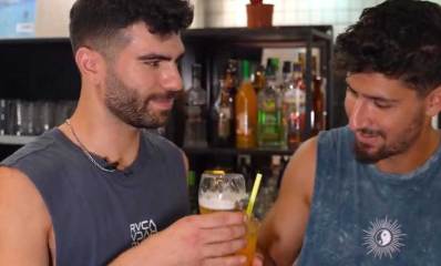 עידן והוד בעלי הבר מרימים כוסות בירה לחיים, לבושים ג'ינס וגופיות כהות על רקע הבר