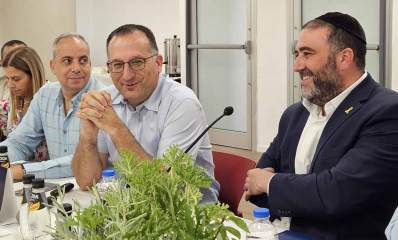 שר הפנים משה ארבל בביקור בעיריית גבעתיים יושב ליד השולחן לבוש חליפה וחולצה לבנה עם סיכה צהובה, סרט החטופים. לידו ראש העיר רן קוניק ועוד שלושה מנהלים. על השולחן לפני שר הפנים עציץ עם צמח ירוק.