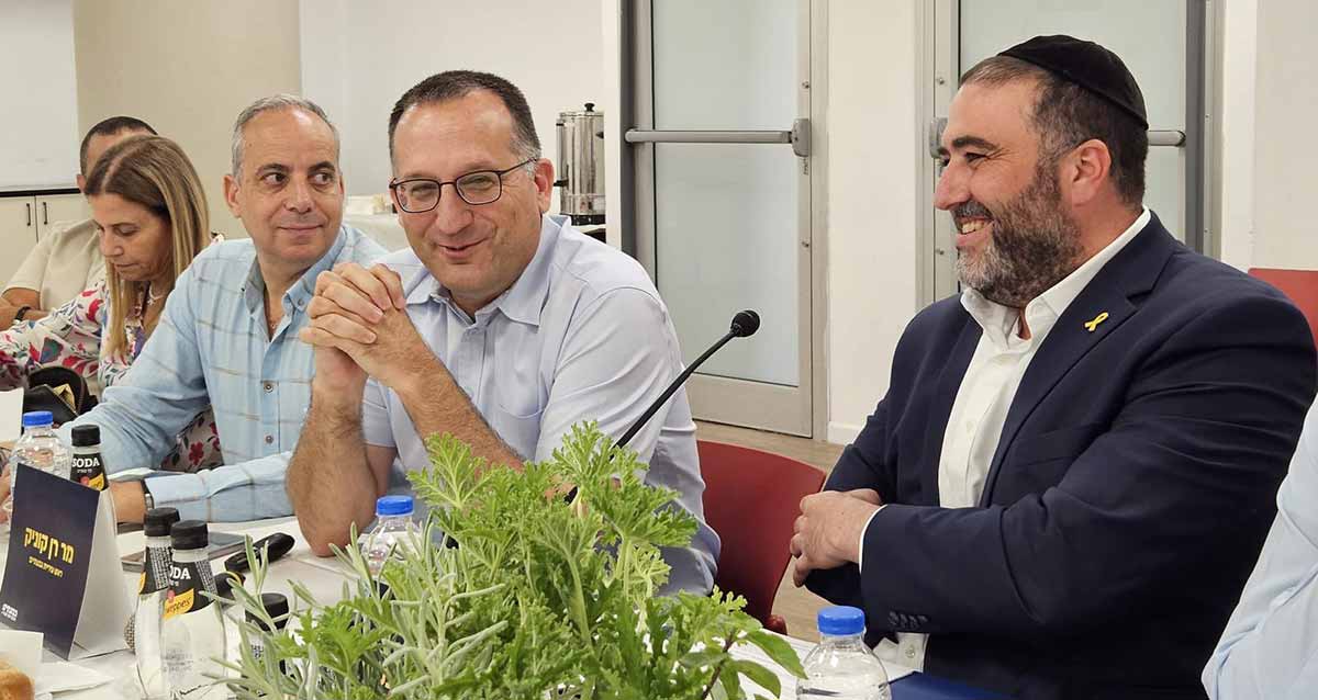 שר הפנים משה ארבל בביקור בעיריית גבעתיים יושב ליד השולחן לבוש חליפה וחולצה לבנה עם סיכה צהובה, סרט החטופים. לידו ראש העיר רן קוניק ועוד שלושה מנהלים. על השולחן לפני שר הפנים עציץ עם צמח ירוק.