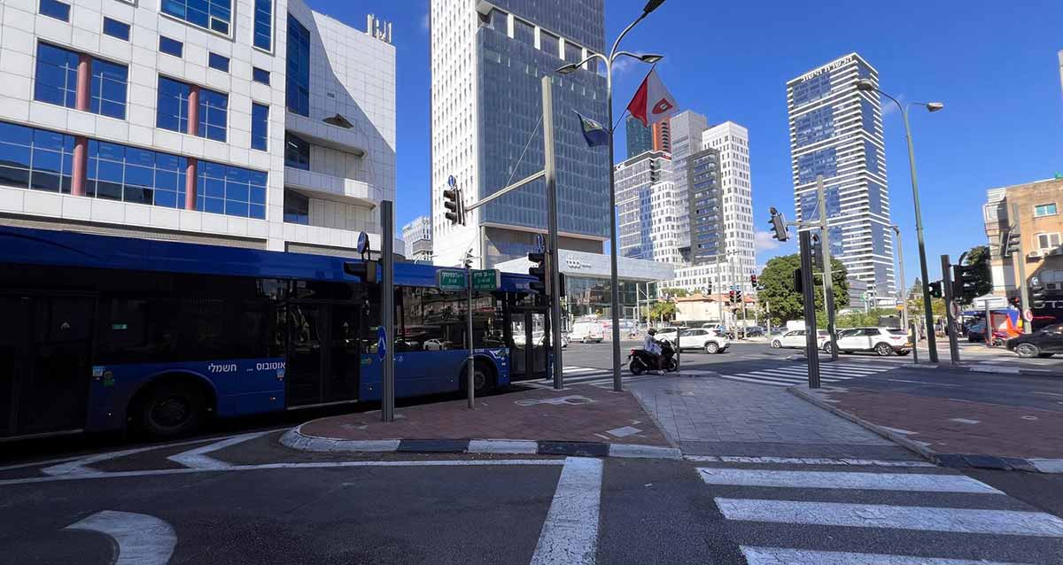 צומת רחוב ז'בוטינסקי ודרך בן גוריון. אוטובוס של דן עומד ליד הרמזור. במרכז שלט עם שמות שני הרחובות בעברית ואנגלית. בצד ימין בניינים רבי קומות. בצד שמאל מעל לאוטובוס בניין של 4 קומות עם חלונות כחולים.