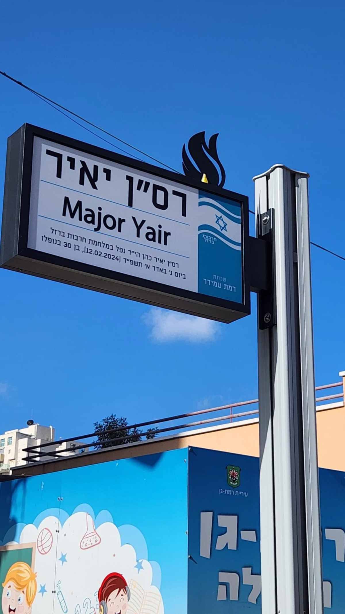 שלט בצבעים כחול ולבן. בצד שמאל על רקע לבן כתוב באותיות שחורות: רס"ן יאיר Major Yair רס"ן יאיר כהן הי"ד נפל במלחמת חרבות ברזל ביום ג' באדר א' תשפ"ד [12.02.2024], בן 30 בנופלו. בחלק הימני של השלט על רקע כחול, למעלה דגל ישראל, מתחתיו כיתוב: חרבות ברזל. בתחתית החלק הכחול כיתוב: שכונת רמת עמידר