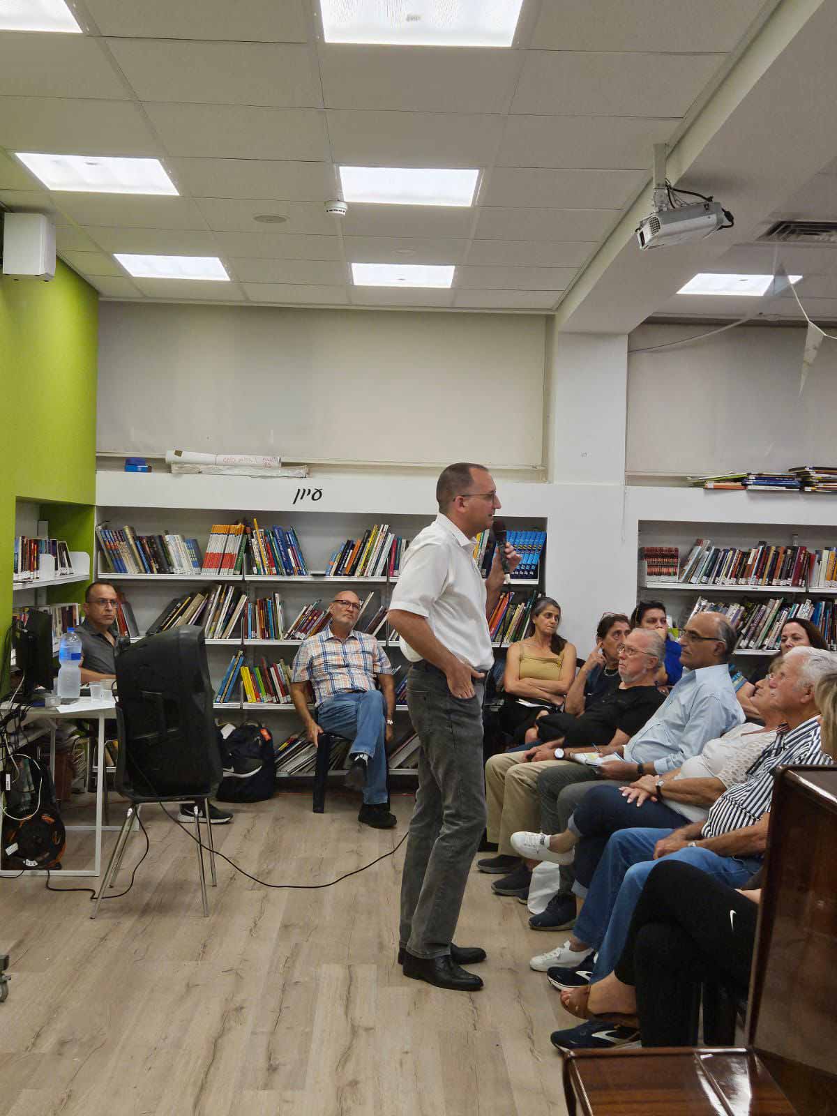 מפגש תושבים עם ראש העיר רן קוניק. ראש העיר עומד באמצע התמונה, לפניו בצד ימין יושבים תושבים בשורות. ברקע קיר של שחר וארוניות עם ספרים צמודות לקיר, ומספר אנשים יושבים לפני הארוניות. ראש העיר אוחז מיקרופון בידו השמאלית, מאחוריו רמקול גדול.