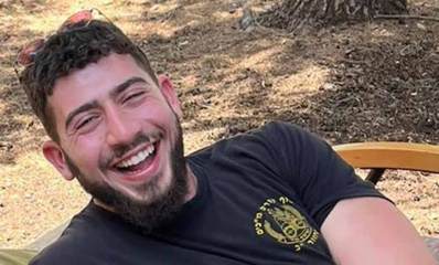 ניב תל צור ז"ל שוכב על מיטה בחורשה. ניב עם חיוך רחב ומשקפי שמש על ראשו. לובש חולצה טישרט שחורה של סוף קורס מכ"ים וסמל היחידה