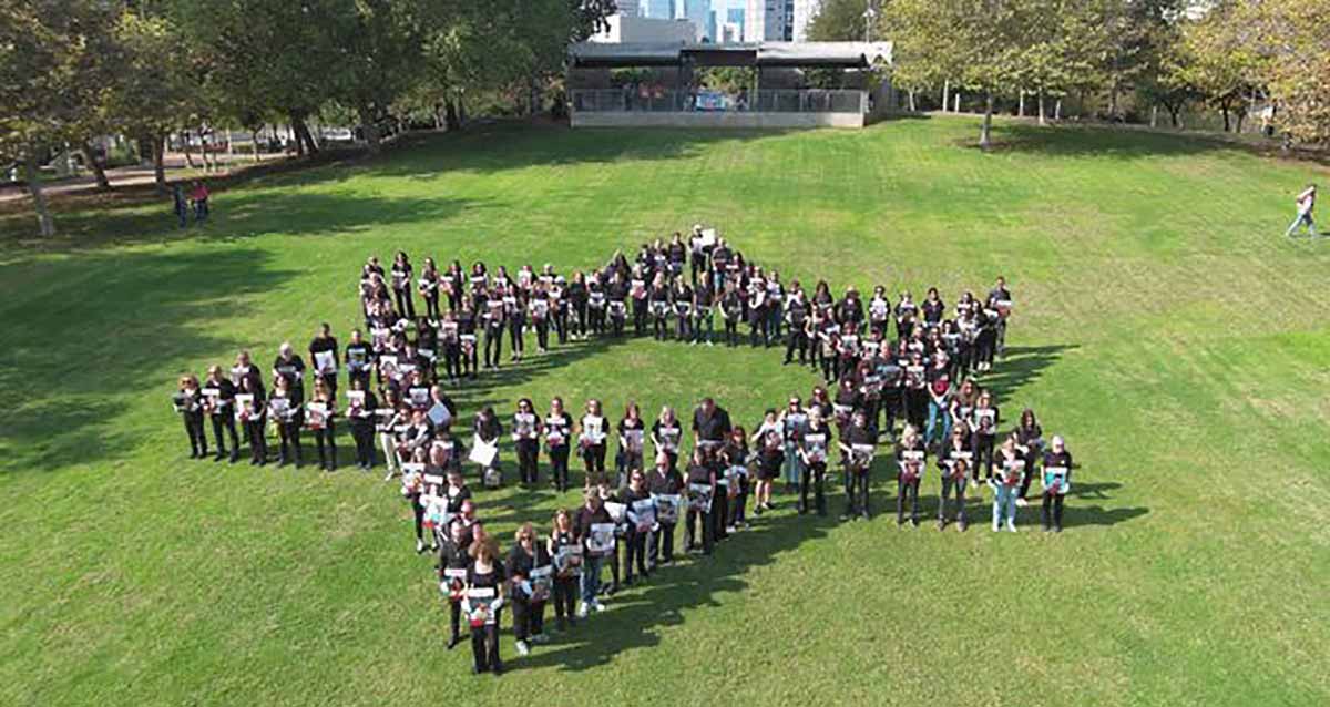 הכנות לציון ה-7 באוקטובר. במרכז התמונה על משטח דשא ירוק וגדול עומדים אנשים בצורת מגן דוד. בידם הם אוחזים בתמונות של חטופים וחללים ברקע מבנה חד קומתי עם חזית של זכוכית ובשני הצדדים עצים.