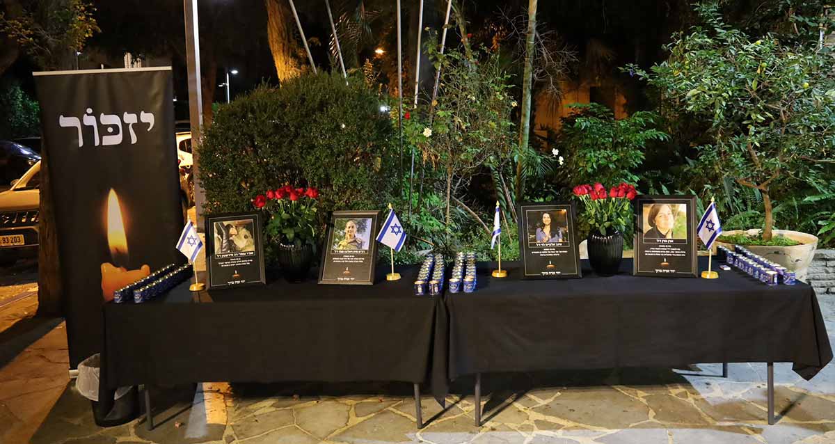 הכנות לציון ה-7 באוקטובר. במרכז התמונה שני שולחנות ארוכים עם מפות שחורות. על השולחנות תמונות של חטופים וחללים, דגלי המדינה ונרות זיכרון. משמאל לשולחנות שלט גדול עם הכיתוב: יזכור, ומתחתיו נר גדול דולק ולהבה גדולה. ברקע עצים.