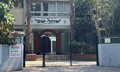 הכניסה לבית הספר "אהל-שם", השערים מהרחוב סגורים. משני צידי שערי הברזל חומת בטון בצבע אפור. בכניסה דלתות עץ חום עם קשת בחלק העליון של הדלת. מעל הדלת כיתובים: בית הספר התיכון העירוני "אהל-שם" "יד לישראל ארצי". משני צידי הכיתוב סמלים. מעל לכיתוב רואים חלק מהקומה הראשונה