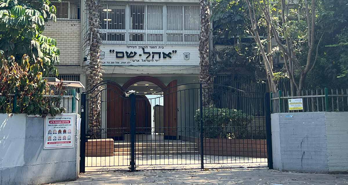 הכניסה לבית הספר "אהל-שם", השערים מהרחוב סגורים. משני צידי שערי הברזל חומת בטון בצבע אפור. בכניסה דלתות עץ חום עם קשת בחלק העליון של הדלת. מעל הדלת כיתובים: בית הספר התיכון העירוני "אהל-שם" "יד לישראל ארצי". משני צידי הכיתוב סמלים. מעל לכיתוב רואים חלק מהקומה הראשונה