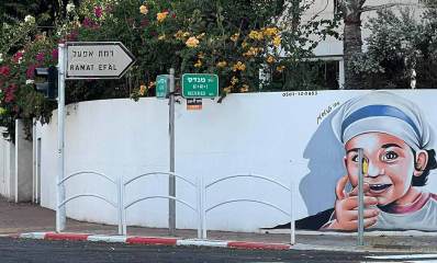 שלט הכוונה לצד ימין לרמת אפעל בפינת רחוב מנדס ורחוב קציר. שלט לבן ועליו בצבע שחור כתוב רמת אפעל בעברית ואנגלית. בחזית חומה של הבית הפינתי, חומת בטון צבועה בלבן ומעוגלת בםינת הרחובות. על החומה מימין ציור גדול של ראש ילדה עם מטפחת ראש לבנה ופס כחול. מעל החומה צמחייה רבה וגדולה וחלק מהבניין הפינתי. בפינת הרחוב שלט רחובות בצבע ירוק מימין רחוב מנדס ומשמאל רחוב קציר.