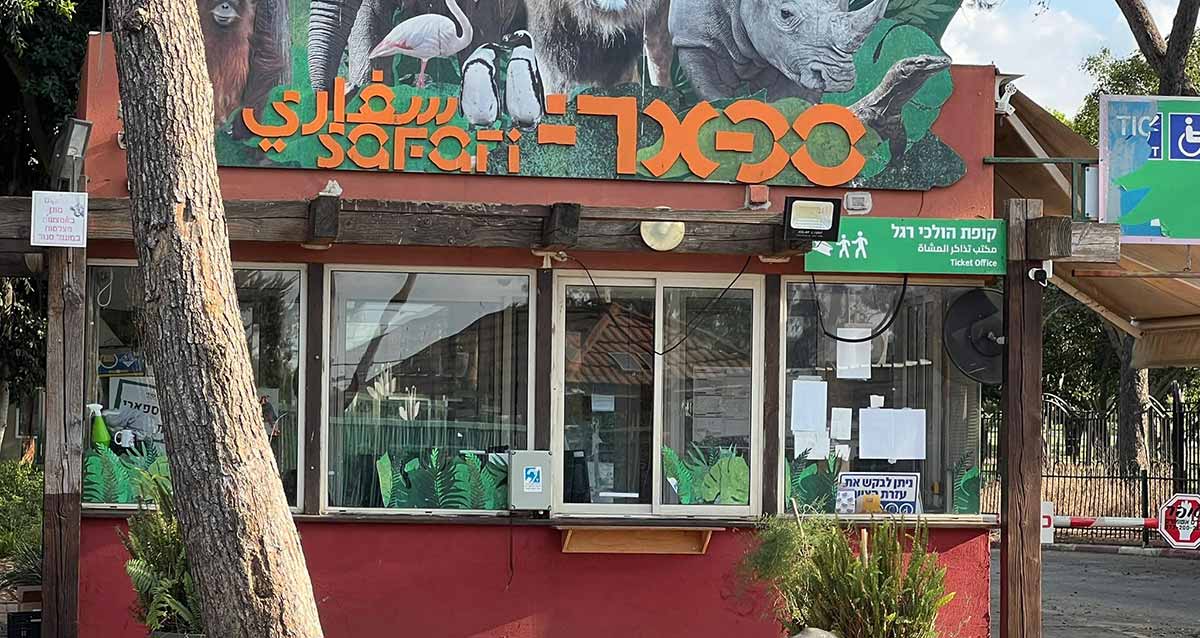 משרדי הספארי ברמת גן וקופות כרטיסים להולכי רגל. בניין צבעוני עם חלונות גדולים לכל רוחבו, צבוע בצבע חום, מעל החלונות שלט לכל רוחב הבניין, ועליו כתוב באותיות גדולות בצבע כתום על רקע ירוק: ספארי בשוש שפות, עברית ערבית ואנגלית. מעל השלט ציורים של חיות, פינגווינים, חסידה, פיל ועוד.בצד שמאל לפני הבניין גזע של עץ. מימין לבניין כביש עם מחסום בצבע אדום ולבן.