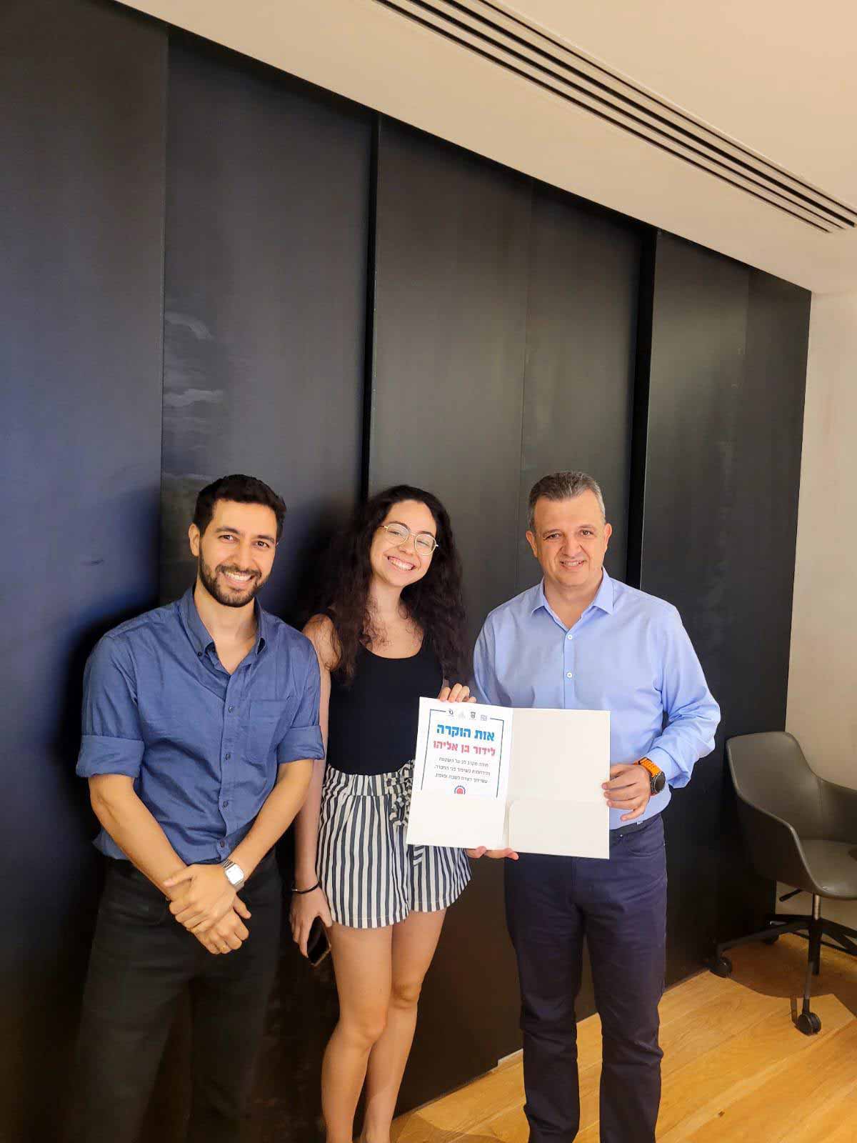 ראש העיר מעניק מלגות לסטודנטים, במשרד על רקע של קיר שחור עומד כרמל שאמה הכהן ראש העיר עם סטודנטית וסטודנט ובידיו תעודת הוקרה של הסטודנטית לידור בן אליהו. הסטודנטית מרכיבה משקפיים ושניהם מחייכים