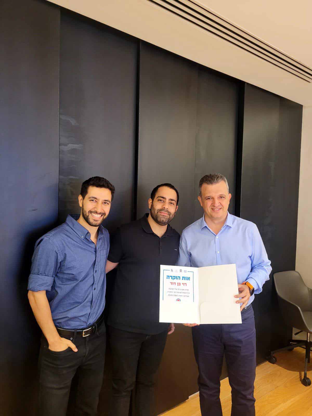 ראש העיר מעניק מלגות לסטודנטים, במשרד על רקע של קיר שחור עומד כרמל שאמה הכהן ראש העיר עם שני סטודנטים ובידיו תעודת הוקרה של אחד הסטודנטים: רוי בן דוד. שני הסטודנטים עם זקנקן