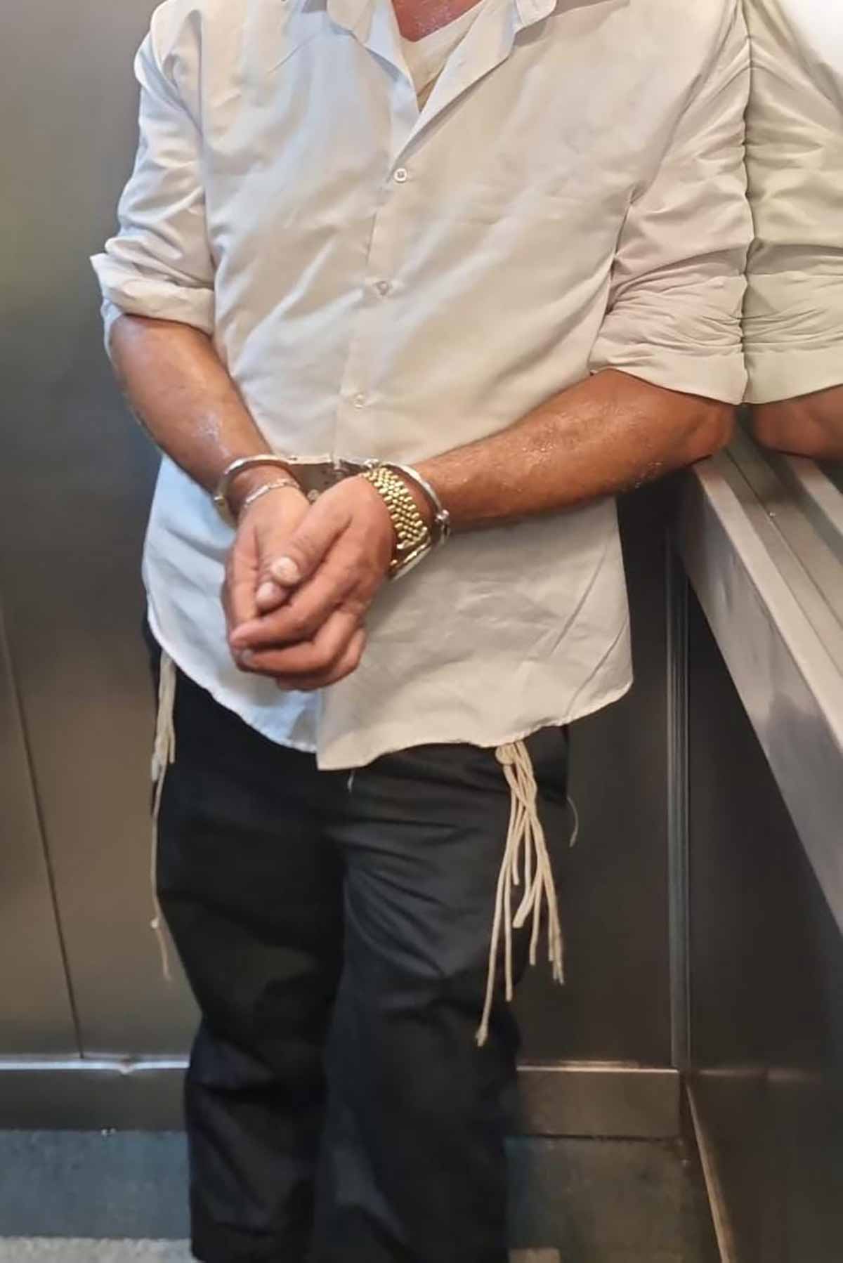 שוהה בלתי חוקי שהתחזה לחרדי, אזוק בידיים, לבוש מכנסיים שחורים וחולצה לבנה ומתחתיה ציציות