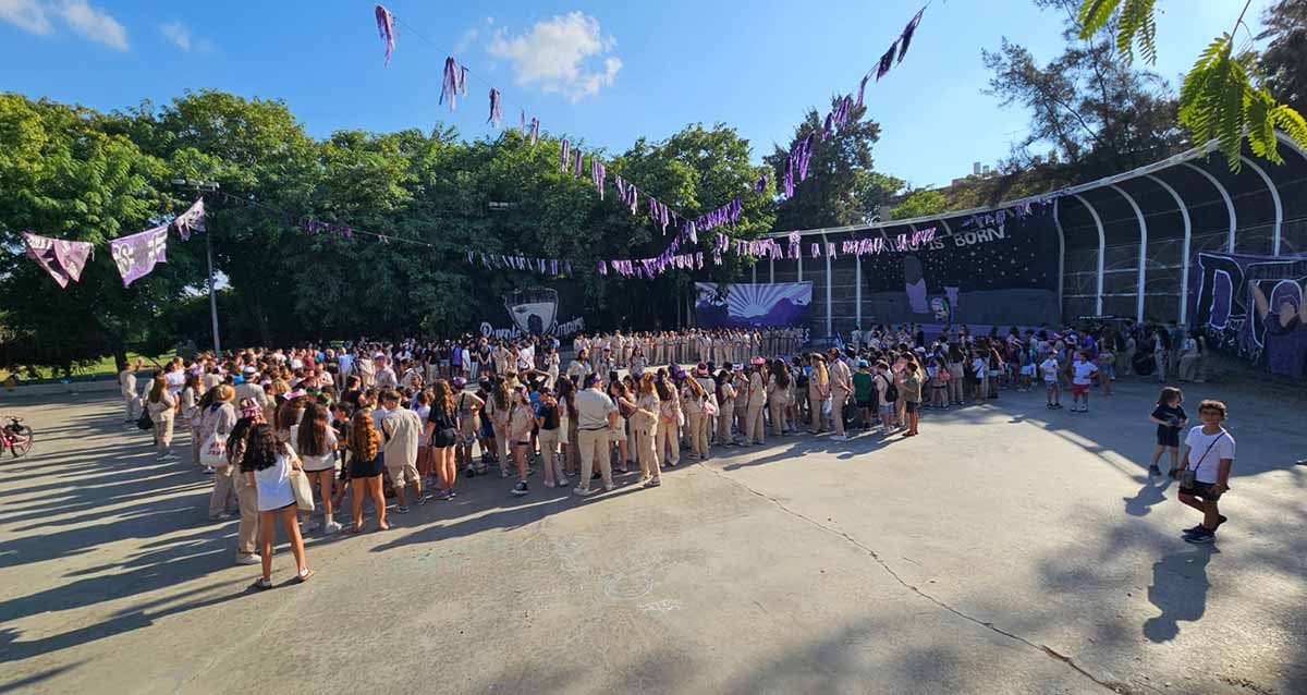 צופים שבט דור גבעתיים, טקס פתיחת השנה החדשה עם 800 חניכים. הצופים עומדים במגרש גדול בשורות ובמדים בצבע חקי. מולם המוני בני המשפחות.מסביב למגרש עצים גדולים ודגלים של הצופים תלויים על חבלים מעל המגרש.