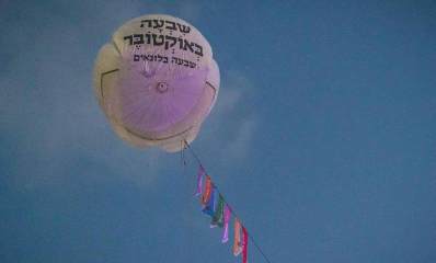 בלון בפארק הלאומי רמת גן, ועליו הכיתוב: שבעה באוקטובר, שבעה בלונאים. הבלון קשור בחבלים למתקן המונח בדשא. על החבל שבעה דגלים צבעונים.
