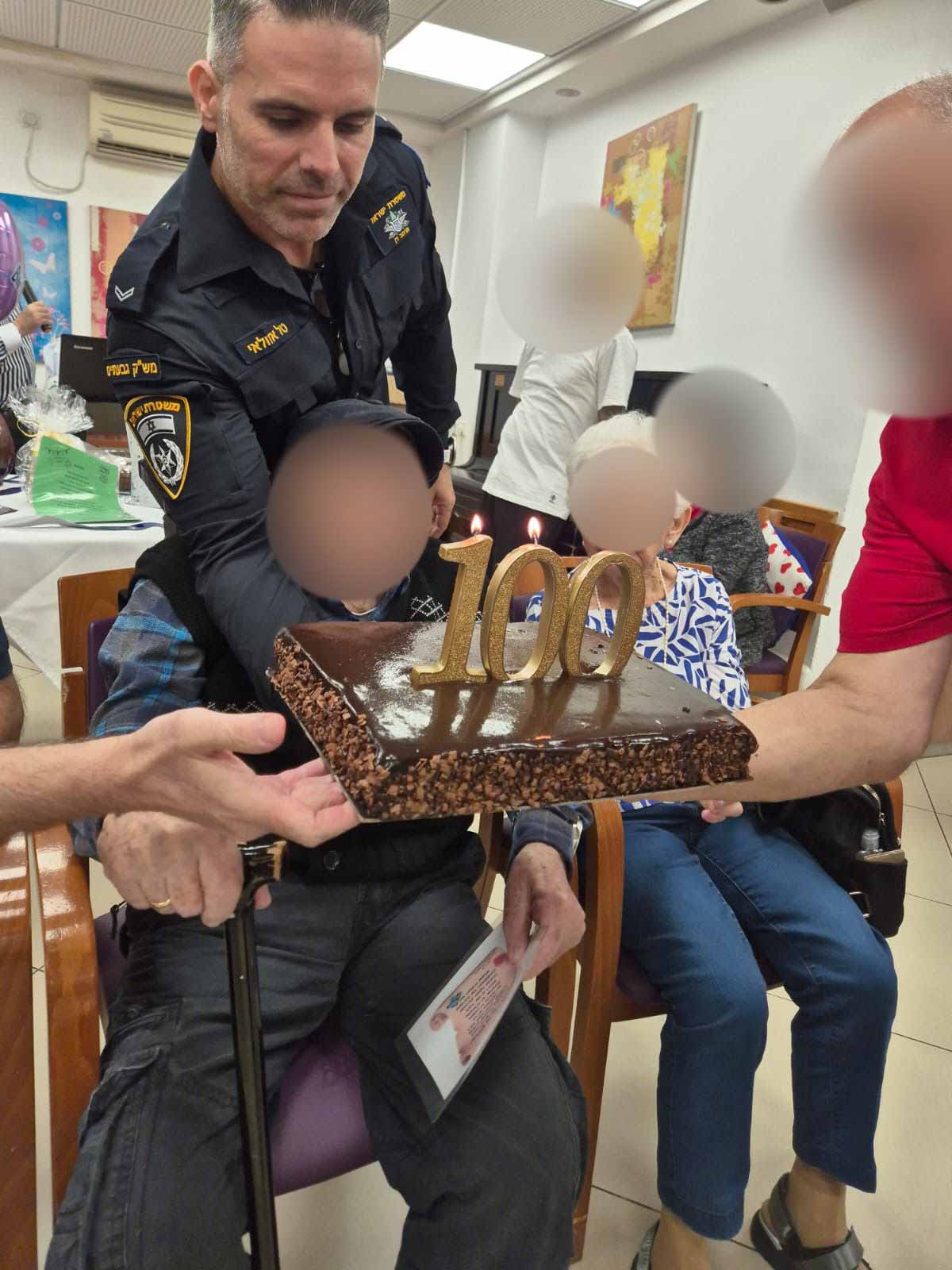 מפקדי ושוטרי תחנת גבעתיים חוגגים 100 לניצול שואה. בן ה-100 יושב על כורסא פניו מוסתרות, וקצין משטרה ואישה מגישים לו עוגת שוקולד ועליה המספר 100. מאחוריו עומד קצין המשטרה, ולידו יושבת אישה על כורסא ופניה מוסתרות.