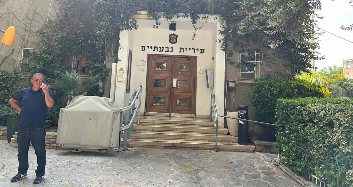 הכניסה לבניין עיריית גבעתיים. ארבע מדרגות מובילות לדלת הכניסה מעץ חום. מעל הדלת הכיתוב: עיריית גבעתיים ומעליו סמל העיר. חזית הבניין מכוסה בצמחים ירוקים. מימין ומשמאל לכניסה גינות של צמחים ירוקים. משמאל לכניסה מוצב מתקן המכוסה בכיסוי לבן. בצד ימין על המדרכה עומד אדם עם טלפון ביד ליד האוזן.