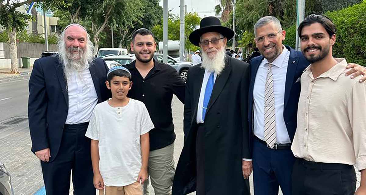 הקפות שניות ברמת גן בסימן החזרת החטופים. עומדים על מדרכה לצילום קבוצתי, הרב יעקב אריאל, הרב יהושע שפירא, וחבר מועצת העיר עו"ד זוהר ישרים וילדיו. ברקע כביש, מכוניות ועצים במדרכות.