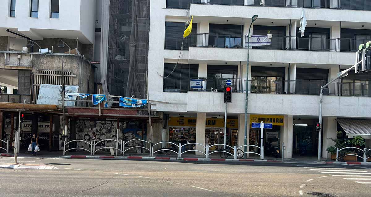 רחוב וייצמן בגבעתיים. מעבר לכביש מדרכה עם גדר מתכת מעוגלת. וברקע בניינים בני ארבע קומות. בקומת הקרקע חנויות. בצד ימין ובשמאל מעברי חצייה להולכי רגל עם רמזורים. אישה ממתינה בצד שמאל לאור ירוק עם שתי שקיות בידיה על מנת לחצות.