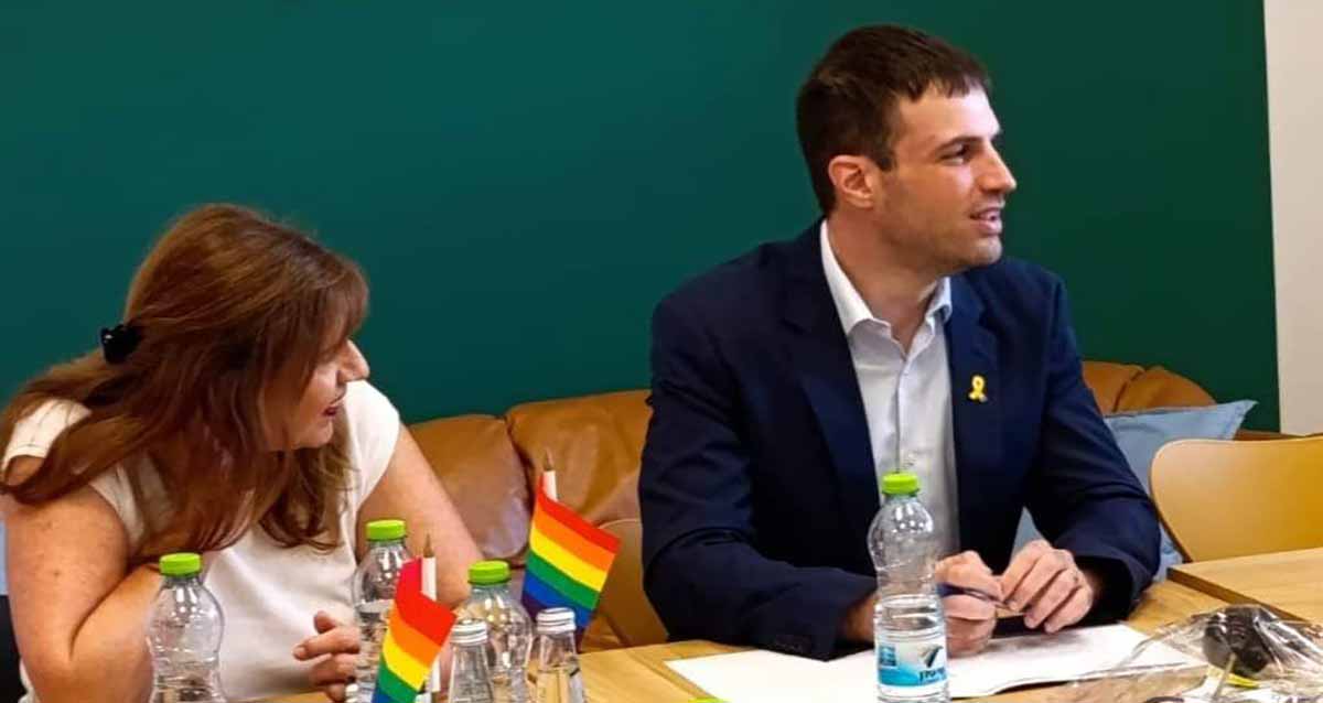 אסף וייס, סגן ראש העיר רמת גן ומחזיק תיק הקהילה הגאה, יושב ליש שולחן ישיבות ומימינו אישה. אסף לבוש חליפה כחולה עם סמל החטופים. על השולחן בקבוקי מים ודגלים של הקהילה הגאה.