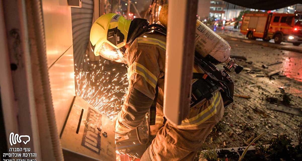 כבאי עם בלון חמצן על גבו, בחליפה וקסדה מנסה לפרוץ למבנה. מוקד שברים ורסיסים על הכביש והמדרכה וכבאית ברקע