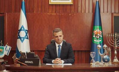 כרמל שאמה הכהן ראש העיר רמת גן, יושב במשרד עם דגל העיר בימין ודגל ישראל בשמאל. על השולחן שלט באנגלית: ISRAEL. מימין לשולחן מדף ועליו חנוכיה. על השולחן מימין פריטים: גלובוס, שעון חול, ועוד.