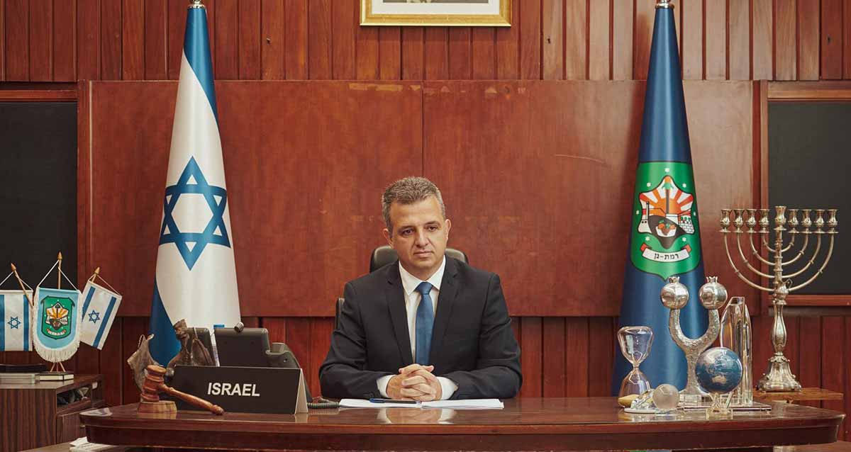 כרמל שאמה הכהן ראש העיר רמת גן, יושב במשרד עם דגל העיר בימין ודגל ישראל בשמאל. על השולחן שלט באנגלית: ISRAEL. מימין לשולחן מדף ועליו חנוכיה. על השולחן מימין פריטים: גלובוס, שעון חול, ועוד.