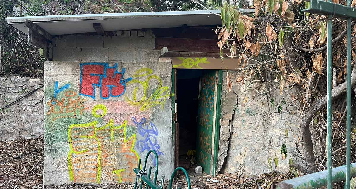 גן הקופים המוזנח ברמת גן, בקתת השומר עשוייה מבלוקים וגגון שטוח. פתח ללא דלת באמצע חזית המבנה. הקיר מקולף בצד ימין. מצד שמאל לדלת יש ציורי קיר בצבעים צהוב, אדום וכחול. עץ יבש בצד שמאל של המבנה.