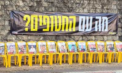 מיצג בדרך יצחק רבין בגבעתיים למען השבת החטופים. ליד החומה שורה של כסאות צהובים ועליהם תמונות החטופים. מעל הכסאות פלקט שחור גדול עם הכיתוב: מה עם החטופים?
