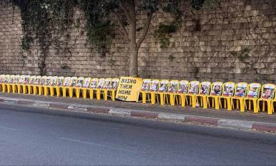 מיצג בדרך יצחק רבין בגבעתיים למען השבת החטופים. ליד החומה שורה של כסאות צהובים ועליהם תמונות החטופים. בין הכסאות פלקט צהוב עם הכיתוב באנגלית: BRING THEM HOME NOW