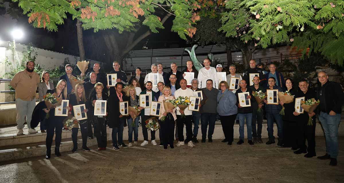 תעודות הוקרה לעסקים בגבעתיים שפועלים למעלה מ-40 שנה. מקבלי התעודות עומדים עם התעודות בשתי שורות, שורה שנייה על משטח גבוה. ברקע עצים עם פריחה.
