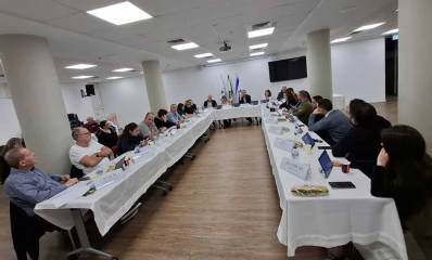 ישיבת מועצת העיר גבעתיים. סביב שולחן בצורת האות ח' המכוסה במפה לבנה, יושבים חברי המועצה. בצד שמאל יושבים אנשים נוספים. על השולחן טאבלטים ובקבוקי שתייה.