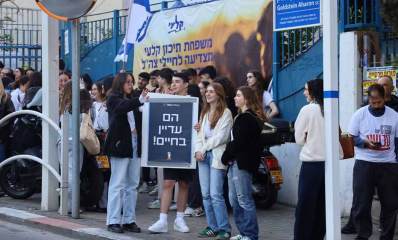 תלמידי גבעתיים מפגינים למען החטופים. תלמידי בית הספר קלעי על המדרכה עם שלטים ודגלים. שלט: הם עדיין בחיים. מאחוריהם פלקט של בית הספר קלעי: משפחת תיכון קלעי מצדיעה לחיילי צה"ל.