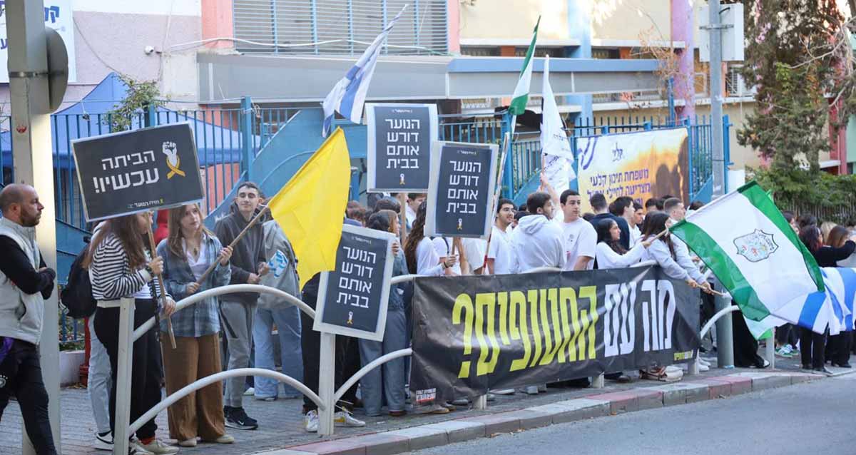 תלמידי גבעתיים מפגינים למען החטופים. תלמידי בית הספר קלעי על המדרכה עם שלטים ודגלים. שלט: הם עדיין בחיים. שלט: הביתה עכשיו! שלט: הנוער דורש אותם בבית. מאחוריהם פלקט של בית הספר קלעי: משפחת תיכון קלעי מצדיעה לחיילי צה