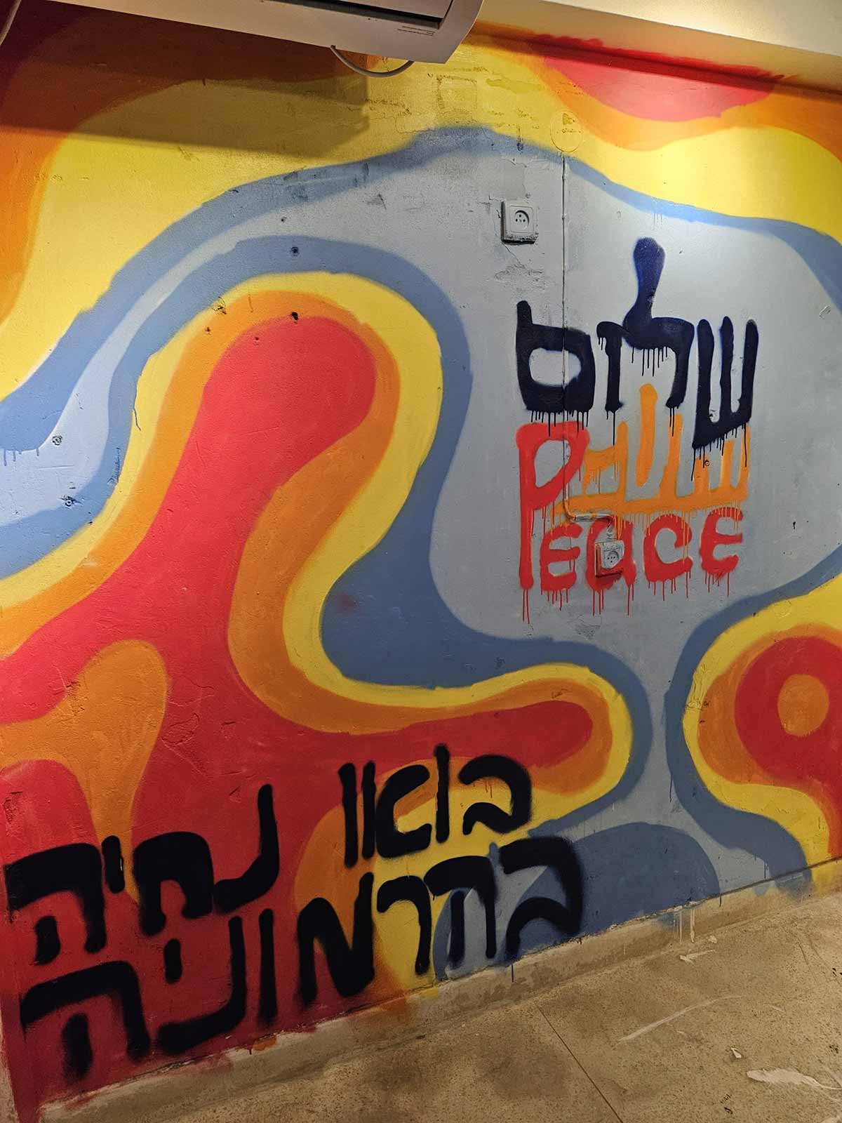 פופ-אפ אמנותי של הנוער על המלחמה. כתובת על הקיר למעלה: שלום בעברית ערבית ואנגלית, ולמטה הכיתוב: בואו נחיה בהרמוניה על רקע של צורות מעוגלות וצבעוניות.