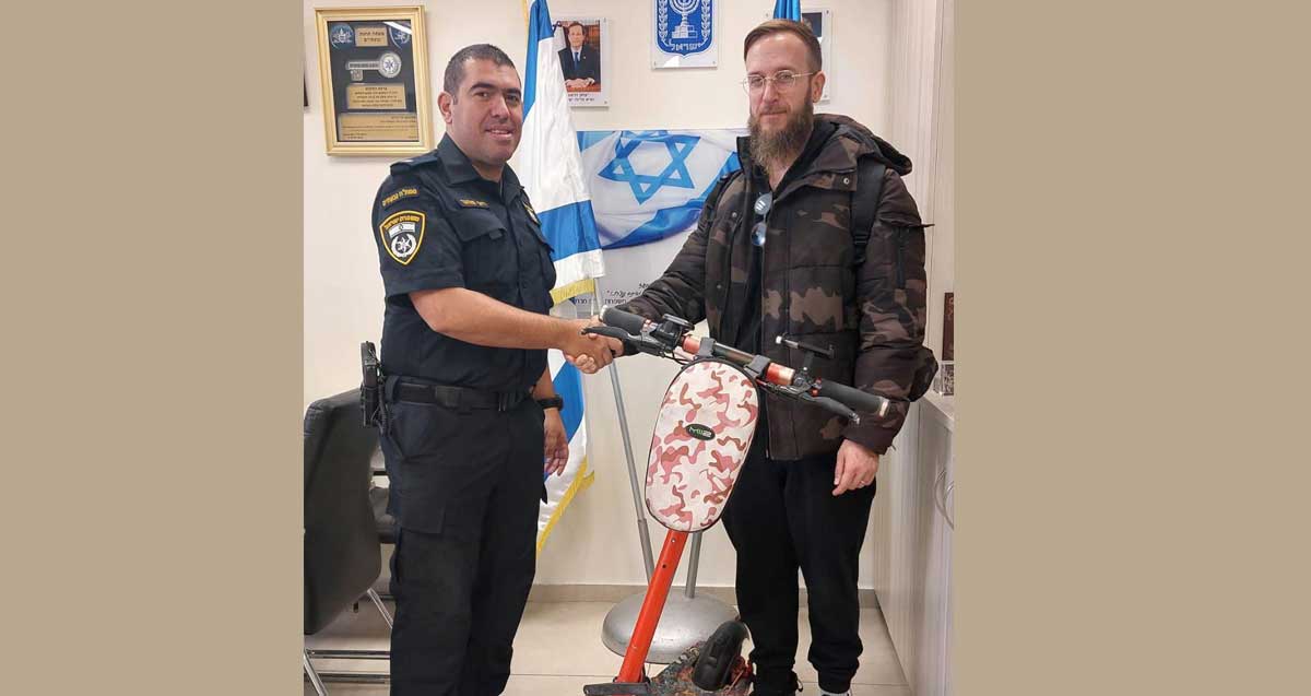 הקורקינט מוחזר לבעליו. השוטר מחזיר את הקורקינט הכתום לבעליו העומד לצידו. ברקע דגלי המדינה, סמל המדינה ותמונות על הקיר.