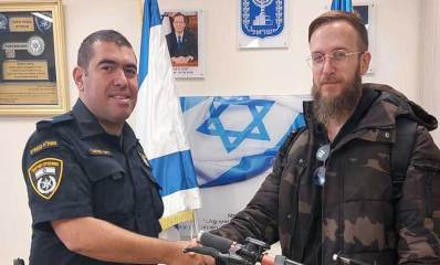 הקורקינט מוחזר לבעליו. השוטר מחזיר את הקורקינט הכתום לבעליו העומד לצידו. ברקע דגלי המדינה, סמל המדינה ותמונות על הקיר.