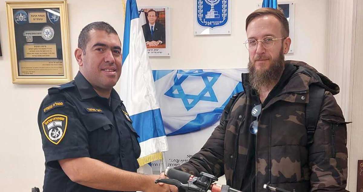 הקורקינט מוחזר לבעליו. השוטר מחזיר את הקורקינט הכתום לבעליו העומד לצידו. ברקע דגלי המדינה, סמל המדינה ותמונות על הקיר.