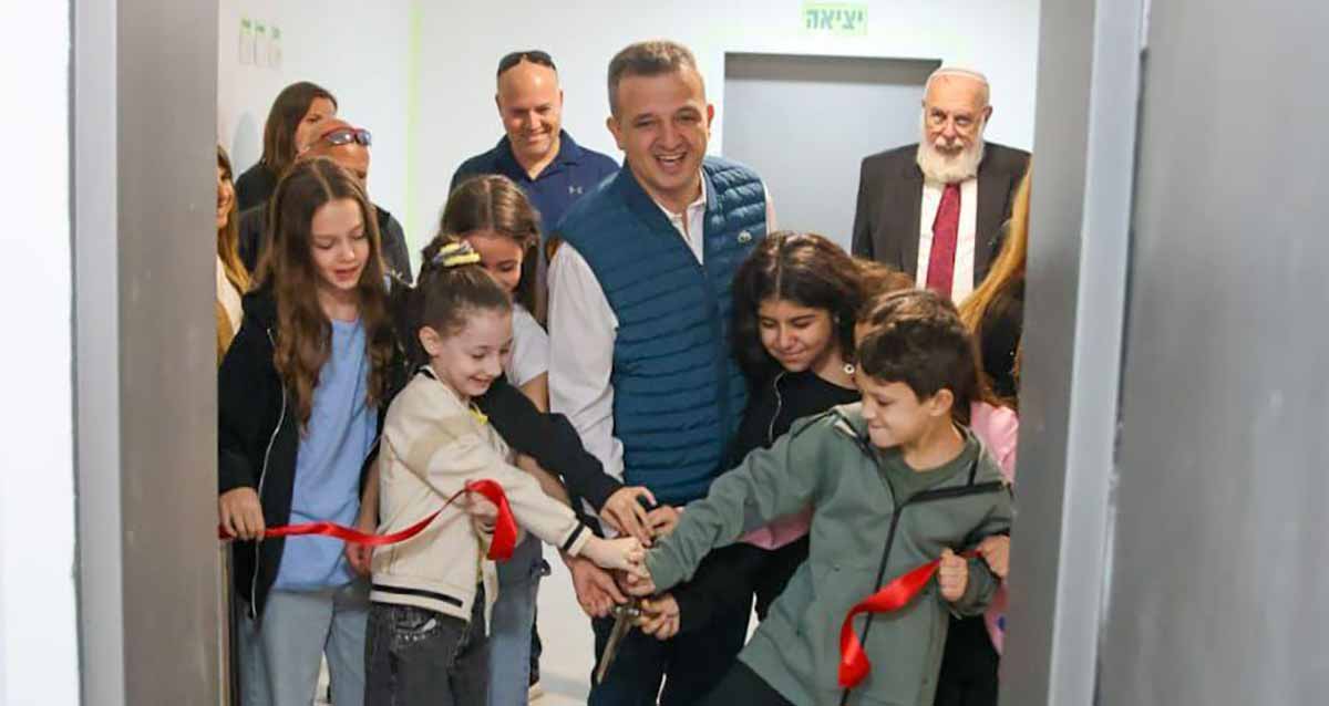 מיגון מוסדות חינוך ומרחבים ציבוריים. ראש העיר עם ילדים מושיטים יד על יד קדימה, בתוך מבנה מוגן נייד, ומאחוריהם מספר מבוגרים.