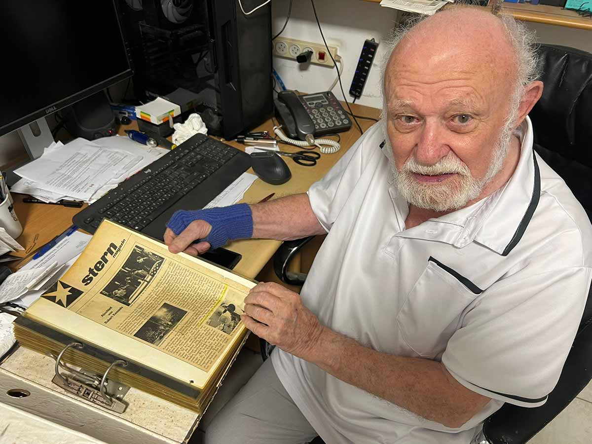 דן הדני, ניצול שואה בן 101, יושב ליד שולחן העבודה וידיו על קלסר עם מסמכים. המסמך המוצג הוא צילום של דף ממגזין בשם: STERN. על השולחן מחשב, מקלדת, עכבר וטלפון.